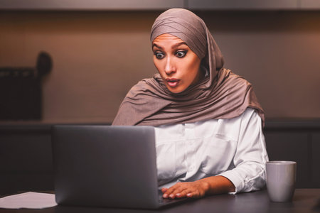 Shocked Muslim Woman Looking At Laptop Browsing Internet In Kitchenの写真素材
