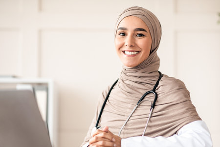 Cheerful doctor in hijab posing at her workdesk in clinicの写真素材
