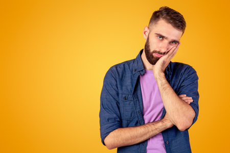 Portrait of bored Caucasian guy leaning on palm and looking at camera on orange studio backgroundの写真素材