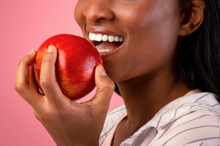Healthy teeth concept. Pretty black woman with beautiful smile biting fresh red apple on pink studio...