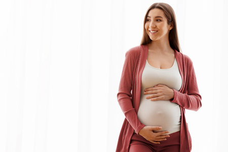 Beautiful Smiling Pregnant Woman Standing Near Window At Home, Touching Her Belly