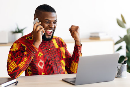 Overjoyed African Businessman In Traditional Clothes Celebrating Success At Workplace In Officeの写真素材