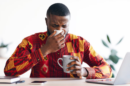 Sick Black Man Sneezing At Workplace In Office, Blowing Nose To Napkinの写真素材