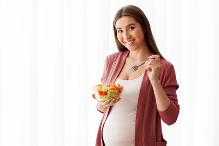 Nutrition During Pregnancy. Smiling Pregnant Lady Eating Fresh Vegetable Salad Near Window