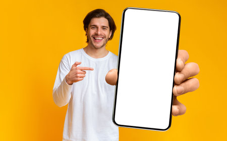 Handsome positive man showing cellphone with white blank screen and pointing on it, mockup, closeup, selective focusの写真素材