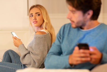 Husband Texting On Cellphone Sitting With Suspicious Wife At Homeの写真素材