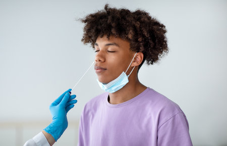 Doctor in rubber gloves making nasal coronavirus PCR test for African American teen, using sterile...