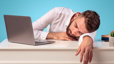 Exhausted young businessman sleeping on his desk, tired of overworking on blue studio background,...