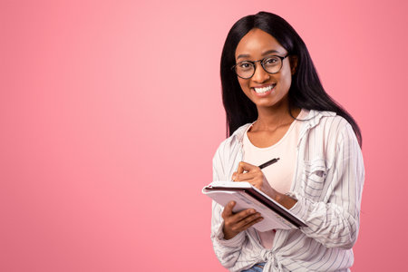 Portrait of intelligent black female student wearing glasses, taking notes in copybook on pink studio backgroundの写真素材