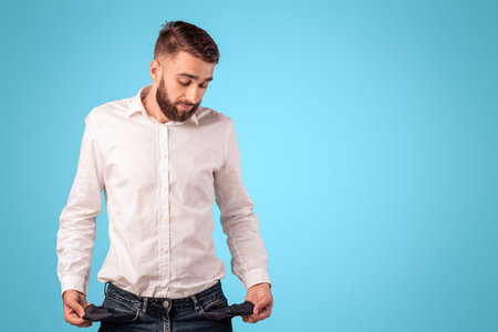 Young bearded man showing empty pockets on blue studio background. Economic depression concept