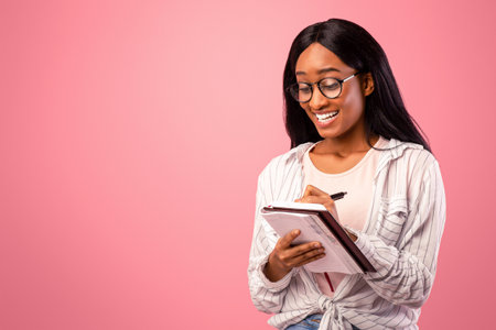 Portrait of smart black lady in glasses writing in notebook on pink studio backgroundの写真素材