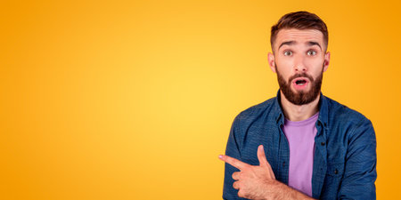 Shocked millennial guy pointing aside at empty space on orange studio background, panorama