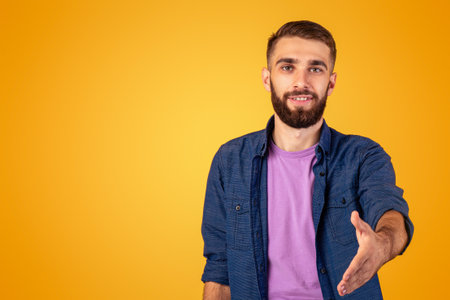 Greeting concept. Smiling Caucasian guy holding out his hand for handshake on orange studio backgroundの写真素材