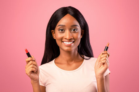 Portrait of happy black lady holding lipstick tubes over pink studio background. Beauty and visage conceptの写真素材