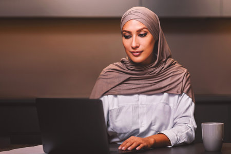 Modern Muslim Woman Working On Laptop Online Sitting In Kitchenの写真素材
