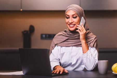 Muslim Lady Talking On Phone Using Laptop In Kitchen Indoorsの写真素材