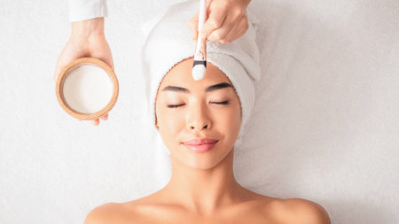 Beauty Treatments. Cosmetologist applying clay mask on asian woman face at spa salon