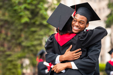 Cheerful black guy hugging his girlfriend while graduation ceremonyの写真素材