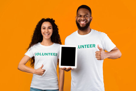 Volunteers Holding Tablet With Empty Screen Gesturing Thumbs-Up, Yellow Background