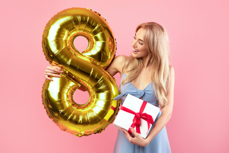 Happy young lady holding number eight golden balloon and bouquet of fresh spring tulips on pink...