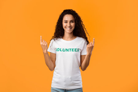 Smiling Volunteer Woman Pointing Fingers Upward Over Yellow Backgroundの写真素材