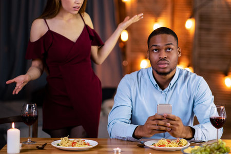 Annoyed woman angry to her black boyfriend stuck in smartphone during dateの写真素材