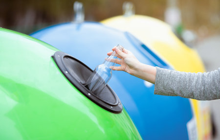 Unrecognizable woman throwing empty glass bottle into green recycle bin outdoors