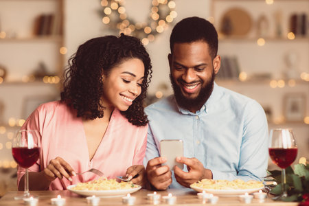 Smiling black man and woman using mobile phone during dinnerの写真素材