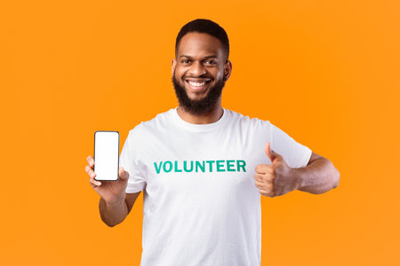 Happy Black Volunteer Showing Phone Screen Gesturing Thumbs-Up, Yellow Background