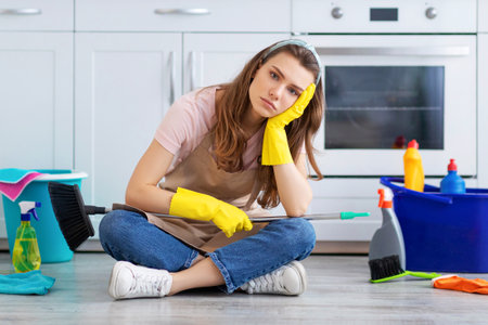 Depressed millennial housewife tired from domestic chores, sitting on kitchen floor surrounded by cleaning suppliesの写真素材