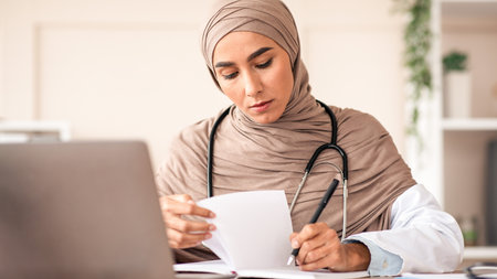 Concentrated young woman doctor in hijab taking notes in clinicの写真素材