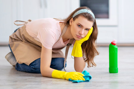 Upset young lady in rubber gloves and apron washing floor at kitchen, disliking house chores,...