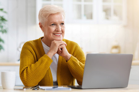 Happy old lady working on laptop at home, kitchen interior