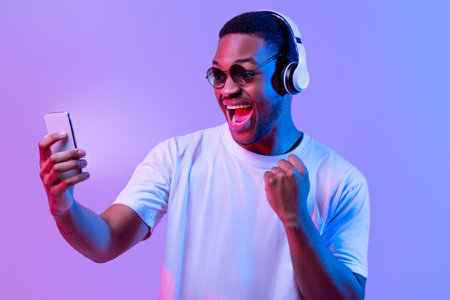 Online Gaming. Excited Black Guy In Headphones Celebrating Success With Smartphone
