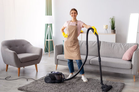 Professional cleaning service. Positive young lady in apron and rubber gloves vacuuming living room of modern houseの写真素材
