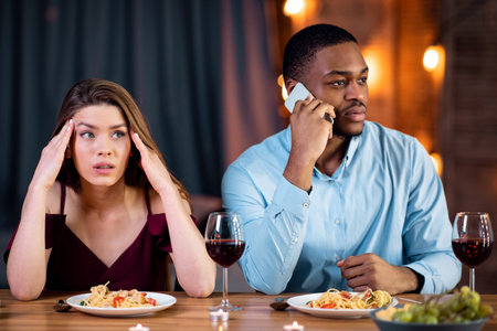 Woman looking unhappy on date while boyfriend talking on cellphone in restaurantの写真素材