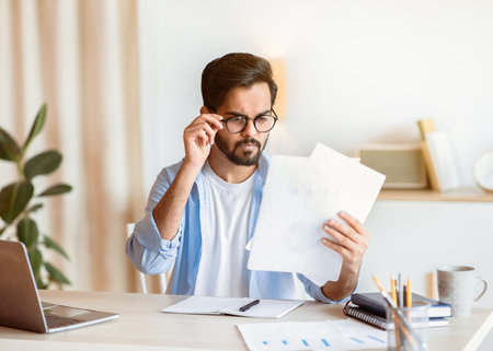 Concerned Arab Businessman In Eyeglasses Reading Financial Documents At Workplace In Office