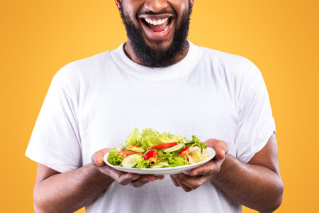 Excited African Man Holding Salad Plate Standing Over Yellow Backgroundの写真素材