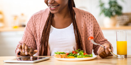 Smiling Young Black Woman Using Digital Tablet While Having Breakfast In Kitchen