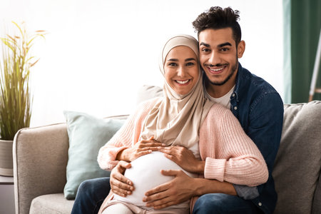 Happy religious muslim couple expecting baby posing on couch at homeの写真素材