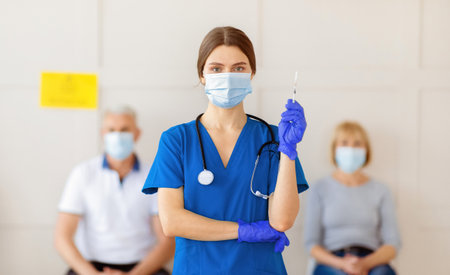 Female doctor in mask and uniform holding syringe with covid-19 vaccine, patients waiting for coronavirus immunizationの写真素材