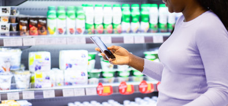 Happy black lady checking her smartphone shopping app while purchasing groceries at dairy section of mall, copy spaceの写真素材