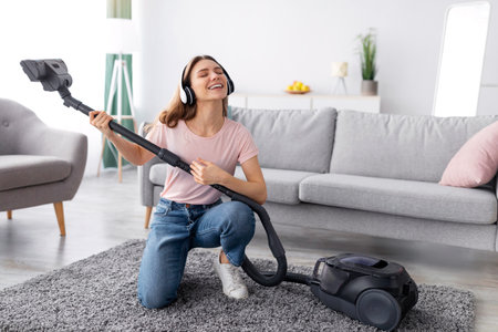 Cleaning is fun. Young housewife listening to music in headphones while vacuuming her apartment, using hoover as guitarの写真素材