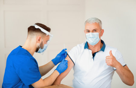 Mature man showing thumb up, receiving covid-19 vaccine injection during his visit to doctor at...