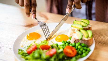 Unrecognizable black female using fork and knife while eating breakfast in kitchen