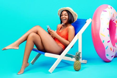 Lovely black woman relaxing in lounge chair, using smartphone, chatting to friend online on beach,...