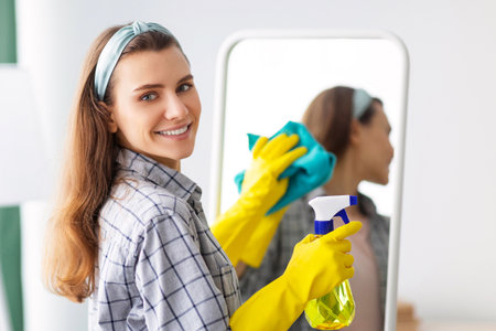 Young Caucasian housewife cleaning mirror, using spray detergent and rag at home. Sanitary service conceptの写真素材