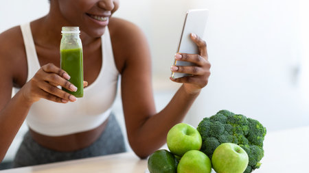 Black Fitness Woman Using Mobile Phone Drinking Smoothie In Kitchenの写真素材