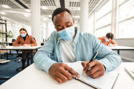 Black guy in medical mask sitting at desk and writingの写真素材