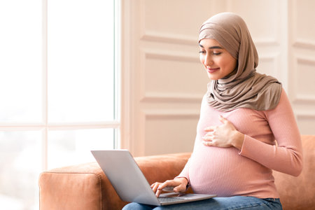Pregnant Muslim Woman Using Laptop Sitting On Couch At Homeの写真素材